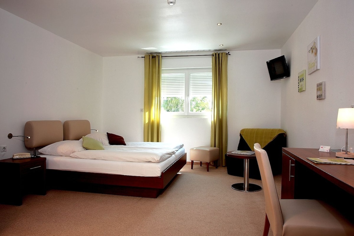 Een hotelkamer met een bed, nachtkastjes, een bureau, een stoel, een aan de muur bevestigde televisie en een raam met gordijnen.