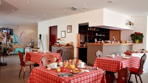 A dining area with red checkered tables and chairs, breakfast items, and a kitchen in the background. - Hôtel les Portes de la Vallée (Turckheim)