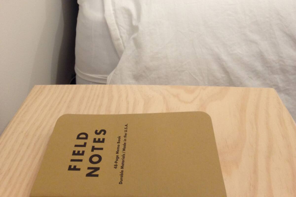 A neatly made bed with white bedding and pillows, a wooden bedside table with a book titled 'FIELD NOTES' on it.