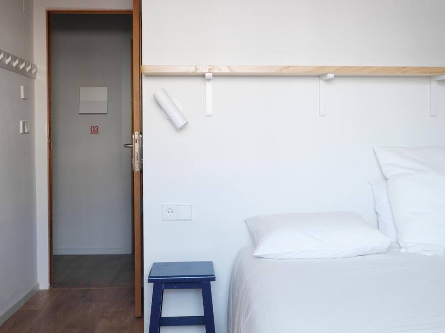 Un dormitorio con cama, un taburete pequeño azul, un estante de madera y una puerta con un número.