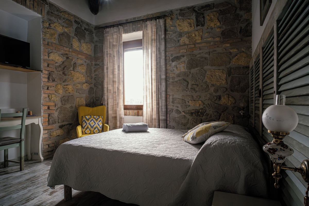 Ein Schlafzimmer mit einer Steinsandsteinwand, einem Bett mit weißer Bettdecke, einem gelben Sessel und einem Nachttisch mit Lampe.