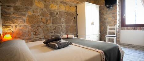 Un dormitorio con pared de piedra, una cama con manta y almohadas, un armario blanco, una silla y una ventana con cortinas.
