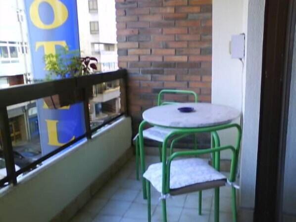 Un balcón con una mesa redonda y dos sillas verdes, un muro de ladrillo y una ventana que refleja edificios.