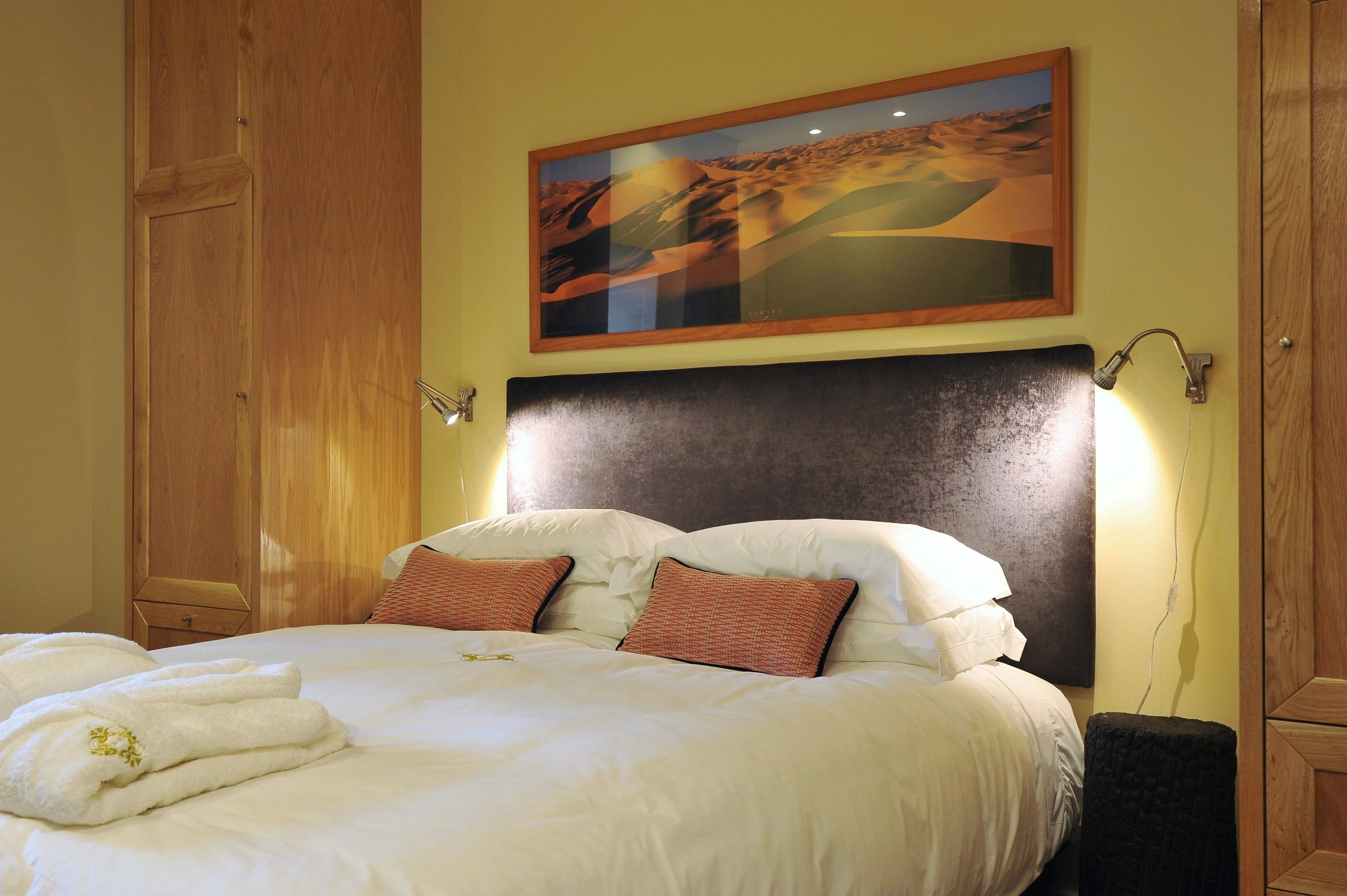 Ein ordentlich bezogenes Bett mit weißen Leinentextilien und zwei Zierkissen, ein hölzernes Kopfende, ein an der Wand befestigtes Gemälde einer Wüstenlandschaft und ein kleiner Nachttisch mit einer Lampe.