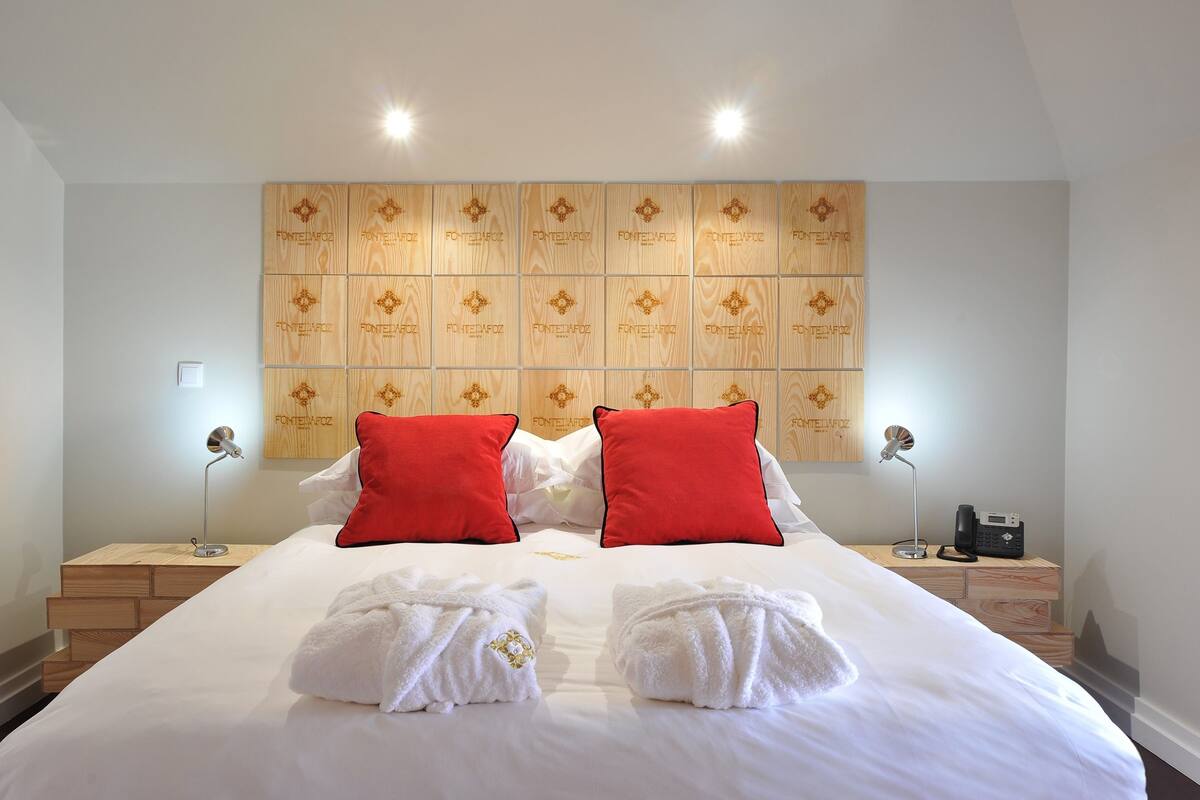 A hotel room with a large bed, two red pillows, white towels, and a wall panel with 'FONTEDAFOZ' branding.