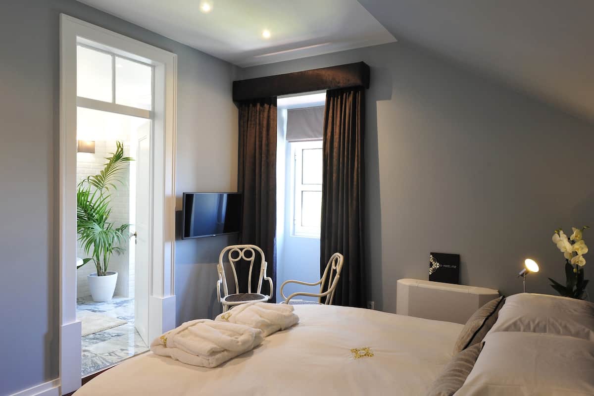 A hotel room with a bed, two chairs, a TV, a bedside table with a lamp, and a window with curtains.