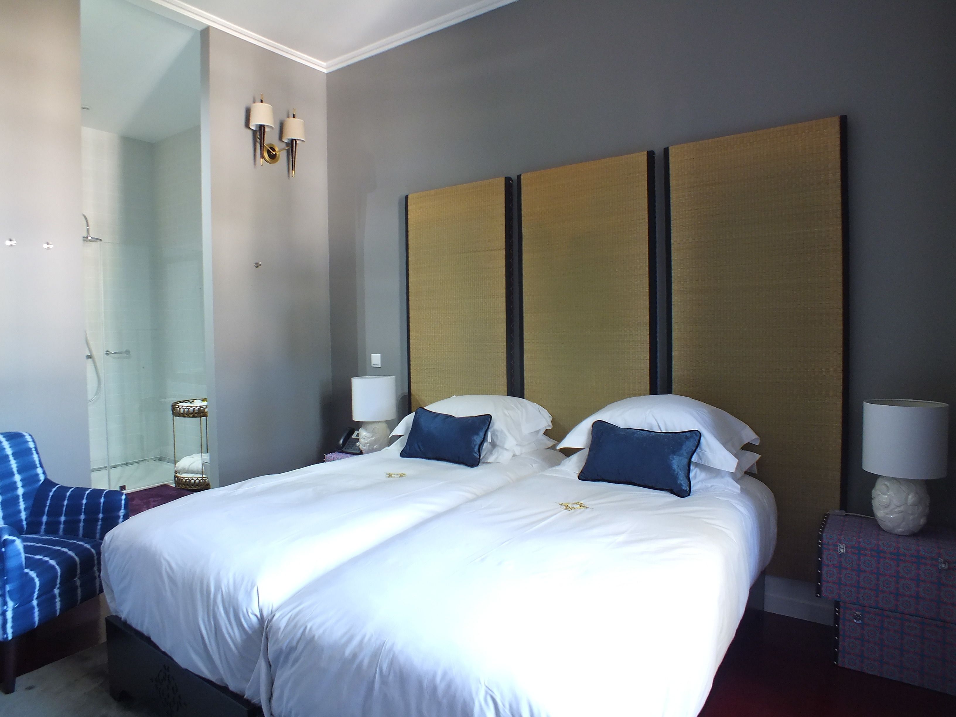 Habitación de hotel con una cama grande, dos mesitas de noche, una silla, un espejo y zona de ducha.