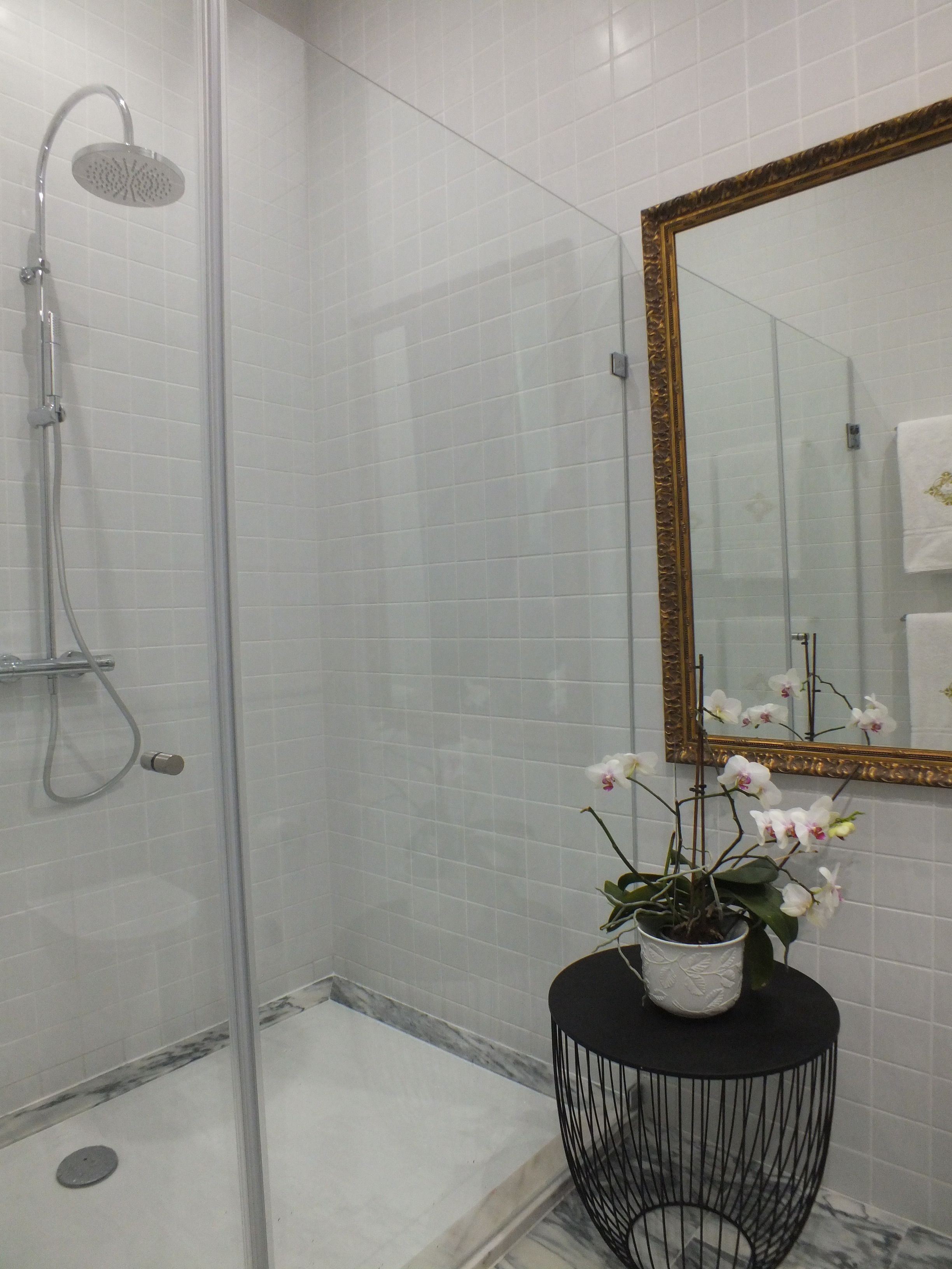 A modern bathroom with a glass shower enclosure, a marble floor, a round black side table with a potted orchid, and a large mirror with a gold frame.