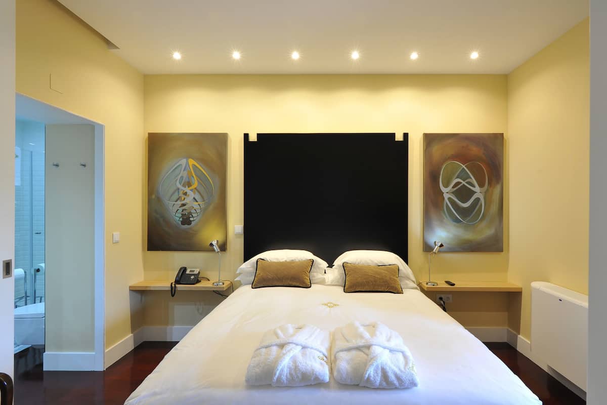 A hotel room with a large bed, two bedside tables with lamps, artwork on the walls, and a bathroom visible through an open door.