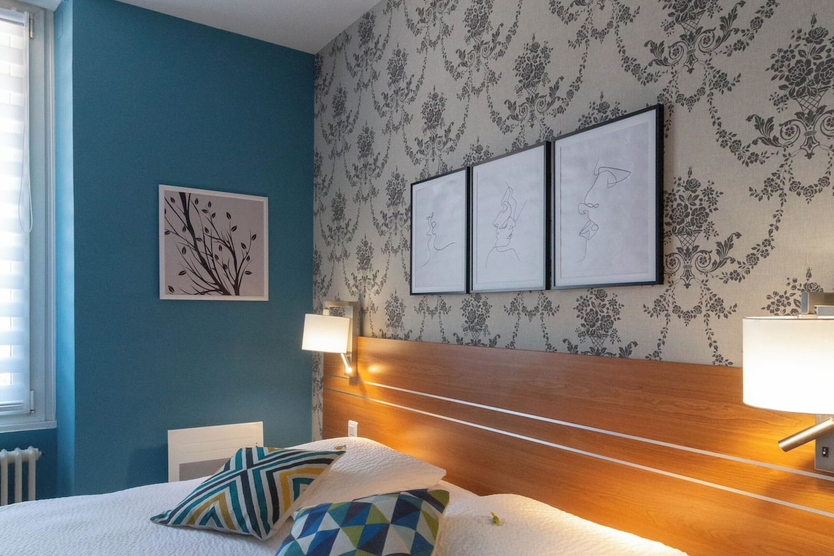 Un dormitorio con cama, una mesita de noche, una pared decorada con cuadros y una ventana con persianas.
