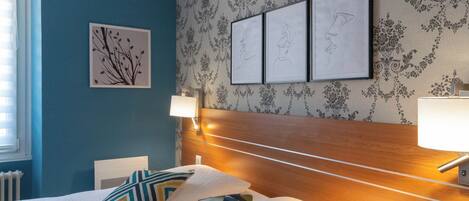 Un dormitorio con cama, una mesita de noche, una pared decorada con cuadros y una ventana con persianas.
