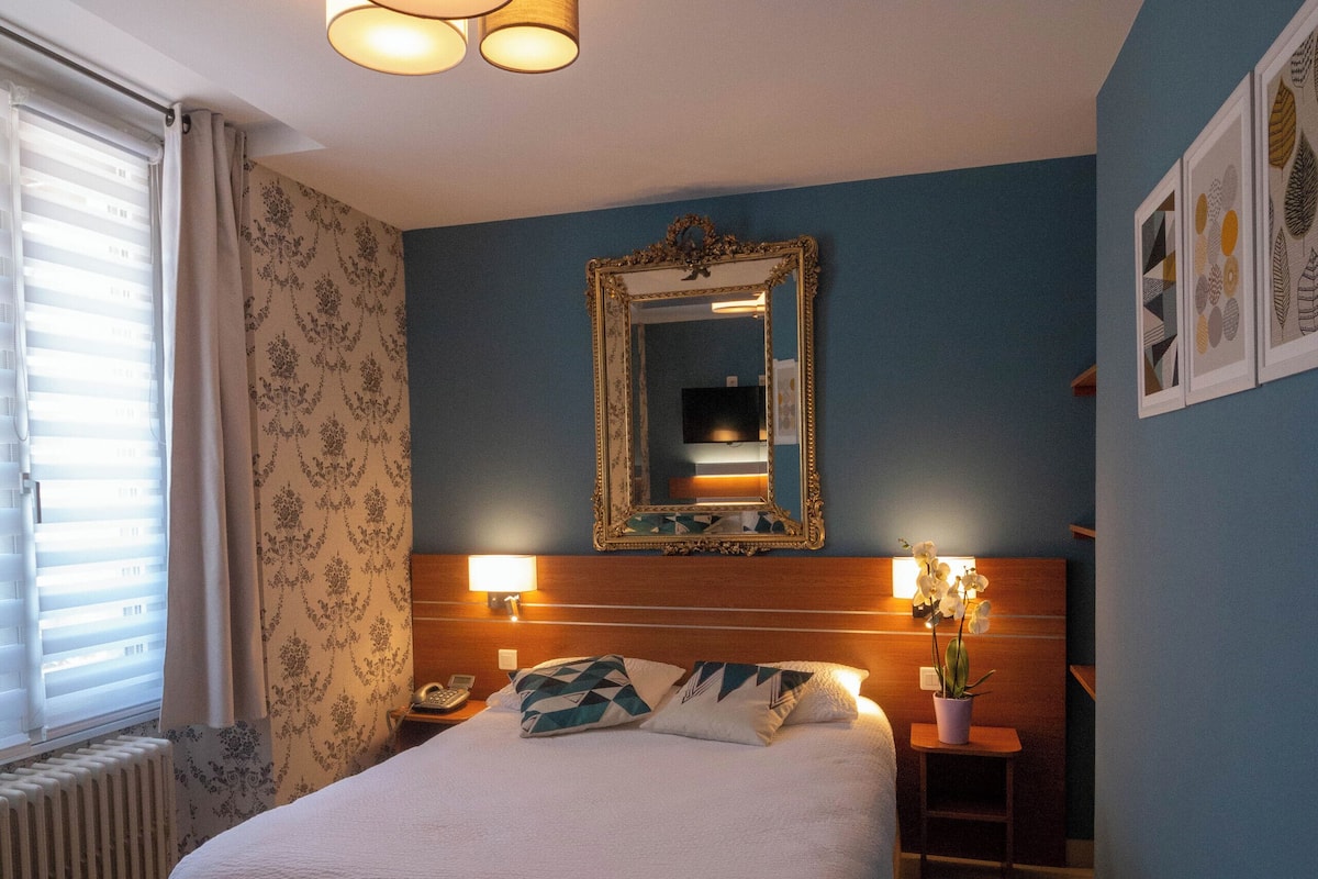 Un dormitorio con una cama grande, cabecera de madera, mesita de noche con lámpara, espejo y ventana con cortinas.