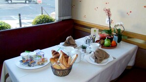 Mesa de café da manhã posta com croissants, pão, frutas e bebidas em um quarto com vista para a rua.
