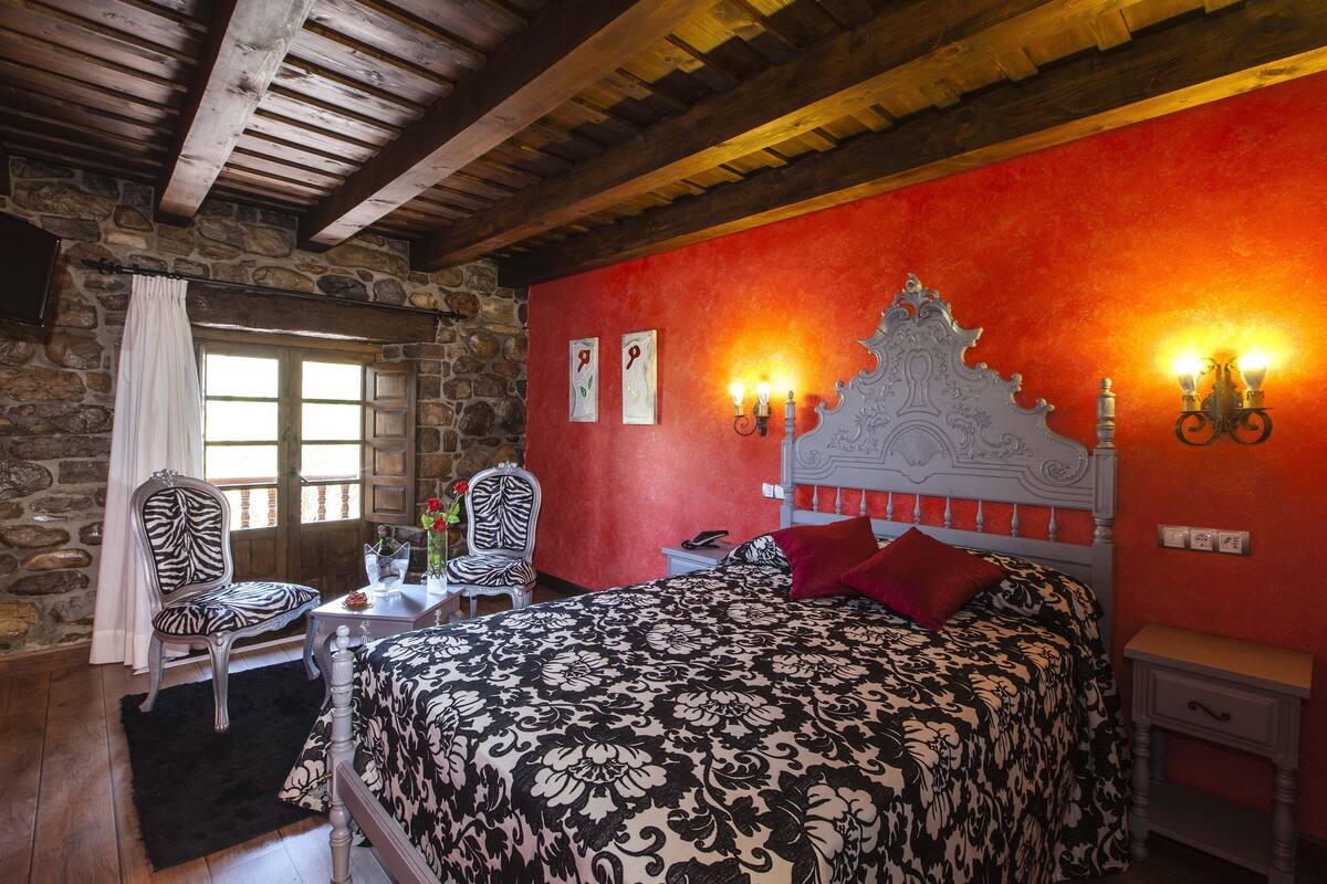 Ein Schlafzimmer mit Steinsturz, einem Bett mit gemustertem Überwurf, einem zebraartig gestreiften Sessel, einer Holzbalkendecke und einem Fenster mit Vorhängen.
