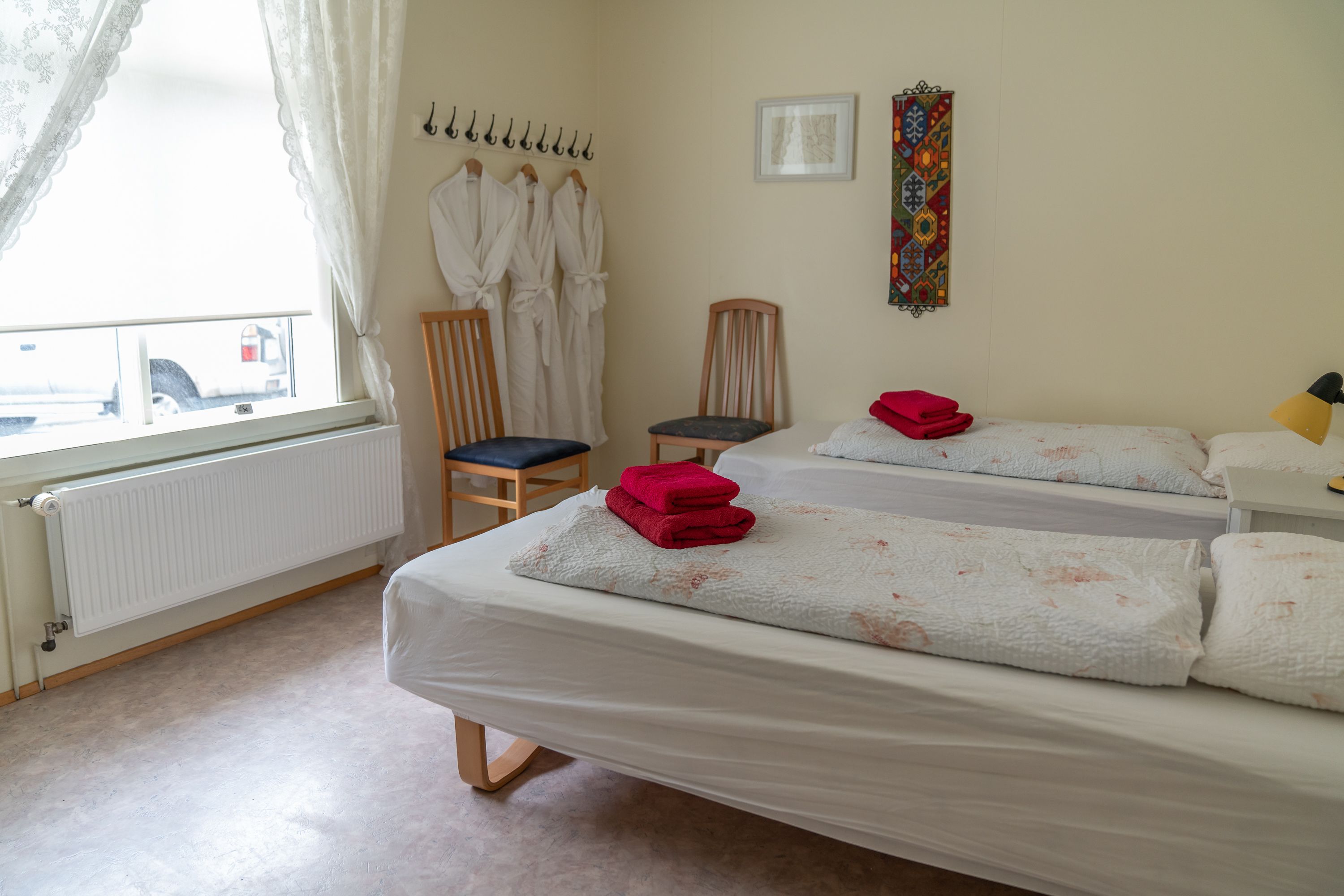 Quarto de hotel com duas camas, uma cadeira, um radiador e uma janela com cortinas.