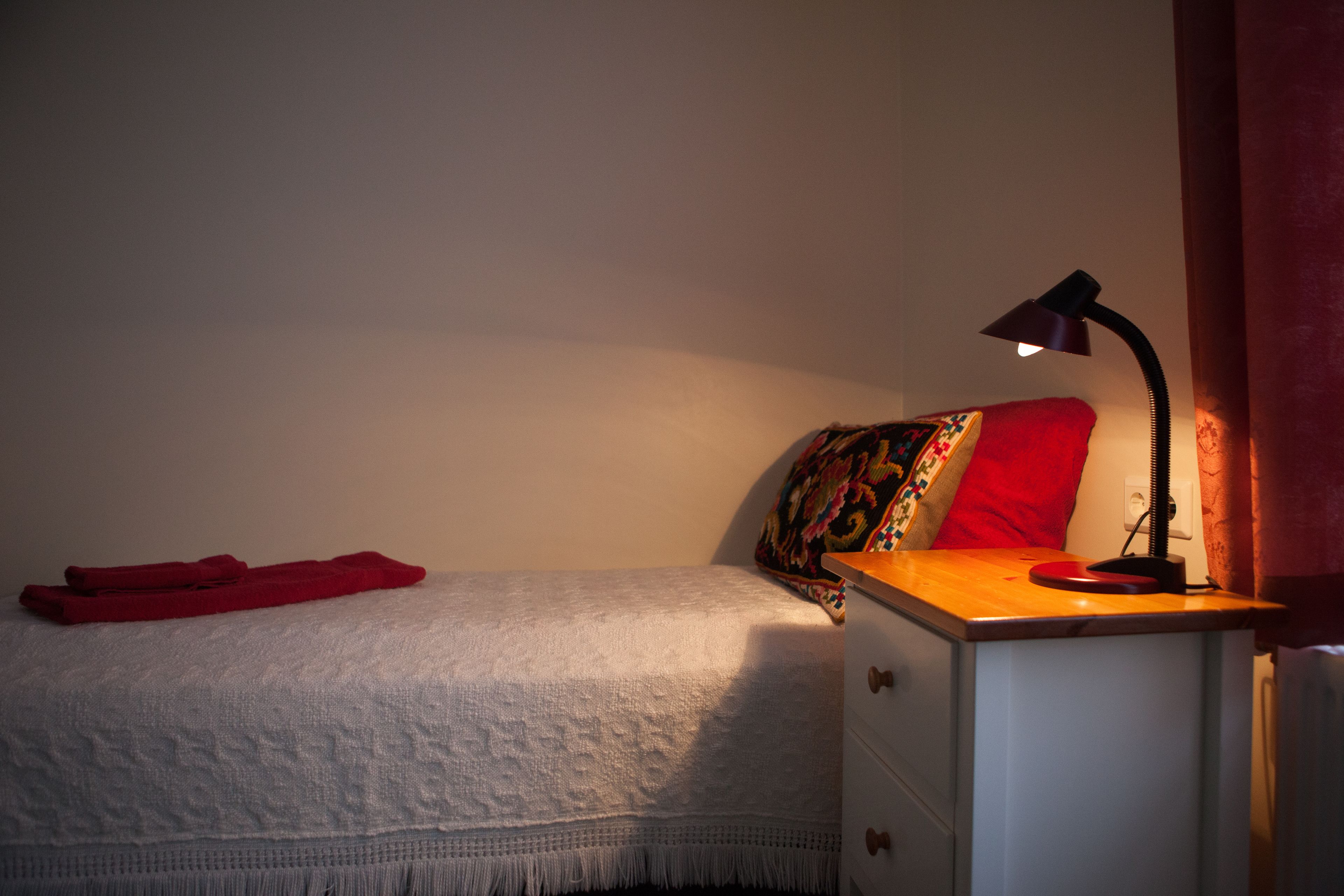 A bedroom with a bed, a bedside table with a lamp, a red towel, and a colorful pillow.
