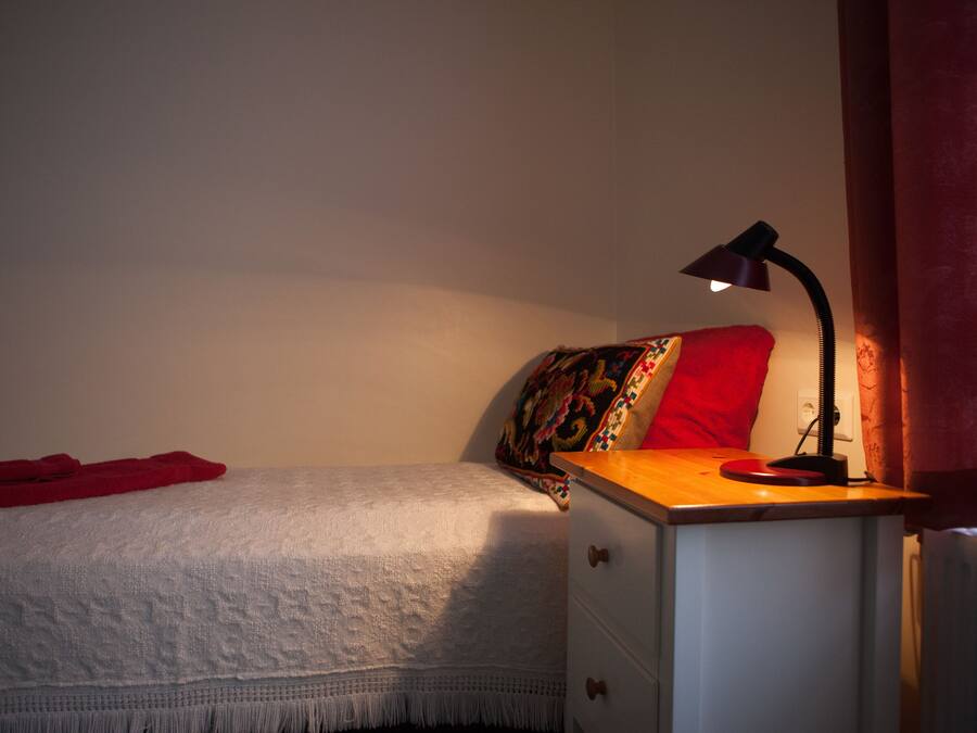 A bedroom with a bed, a bedside table with a lamp, a red towel, and a colorful pillow.