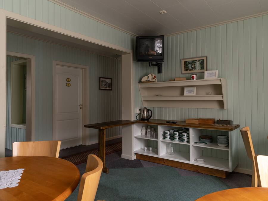 Ein gemütliches Zimmer mit Holztisch, Stühlen, einem an der Wand befestigten Fernseher und einem Regal mit verschiedenen Gegenständen.
