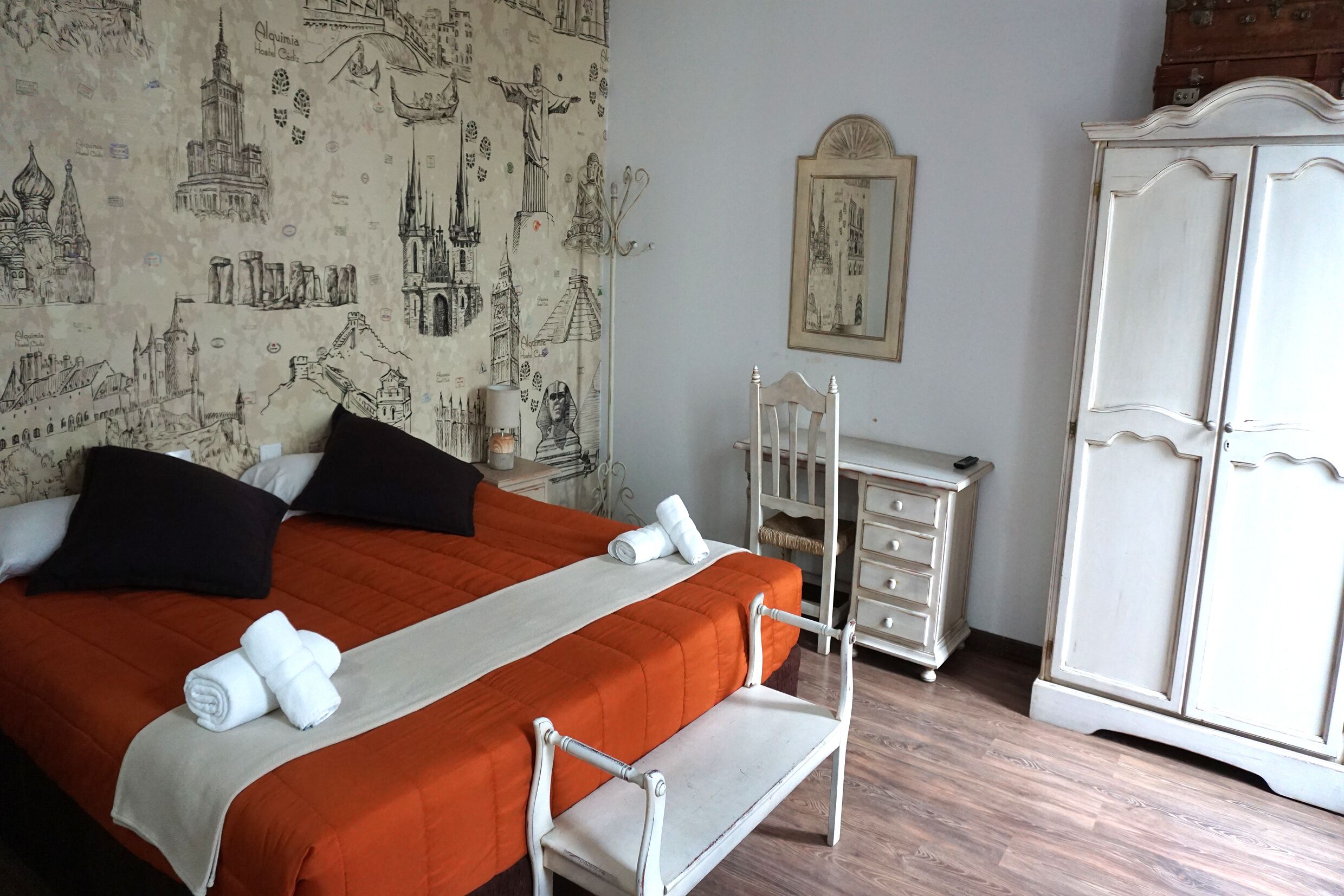 A hotel room with a bed, a nightstand, a chair, a wardrobe, and a wall with a cityscape pattern.