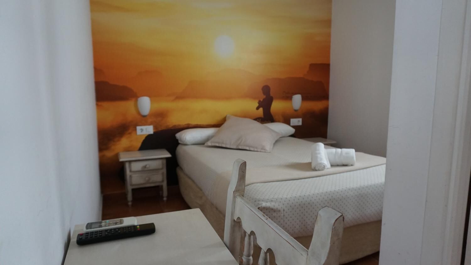 A hotel room with a bed, a nightstand, a remote control, and a mural of a sunset scene.