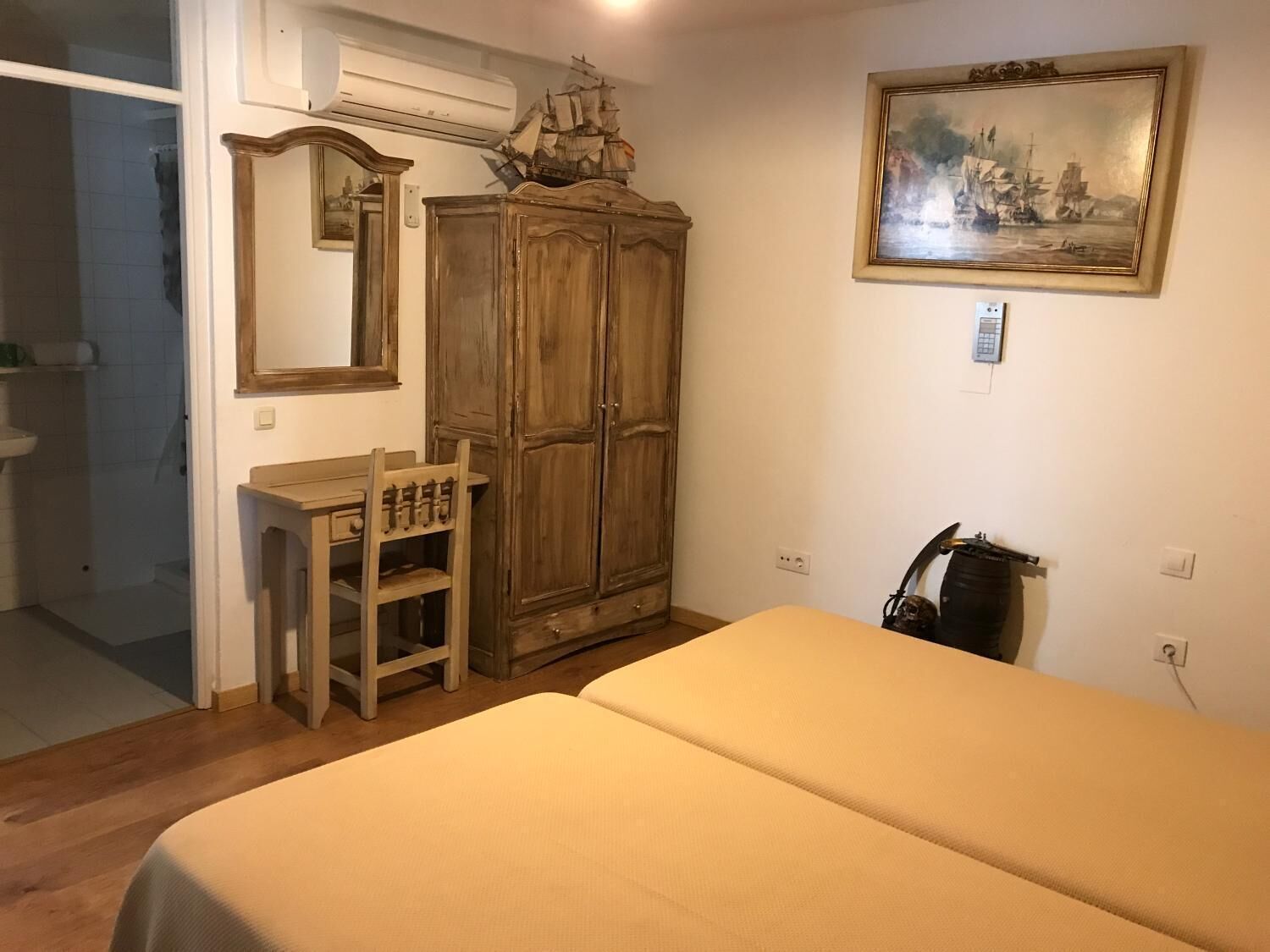 A bedroom with a wooden wardrobe, a desk with a chair, a bed, and a painting on the wall.