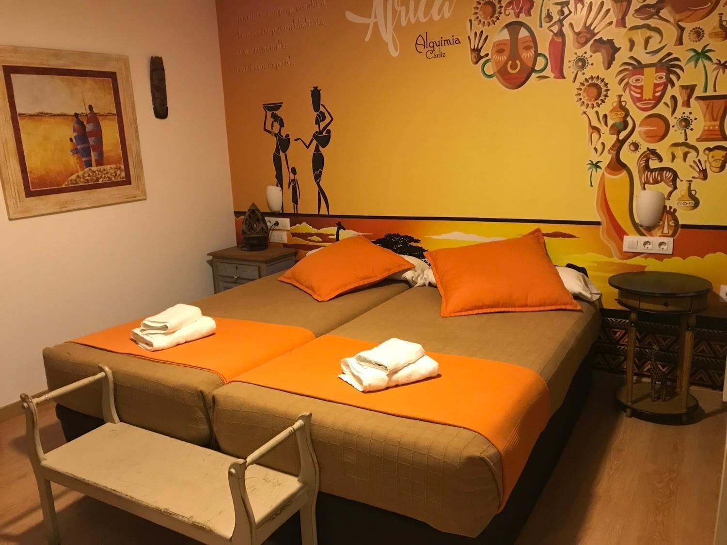 A hotel room with two beds, orange bedding, a small table, a chair, and a wall mural with African cultural motifs.