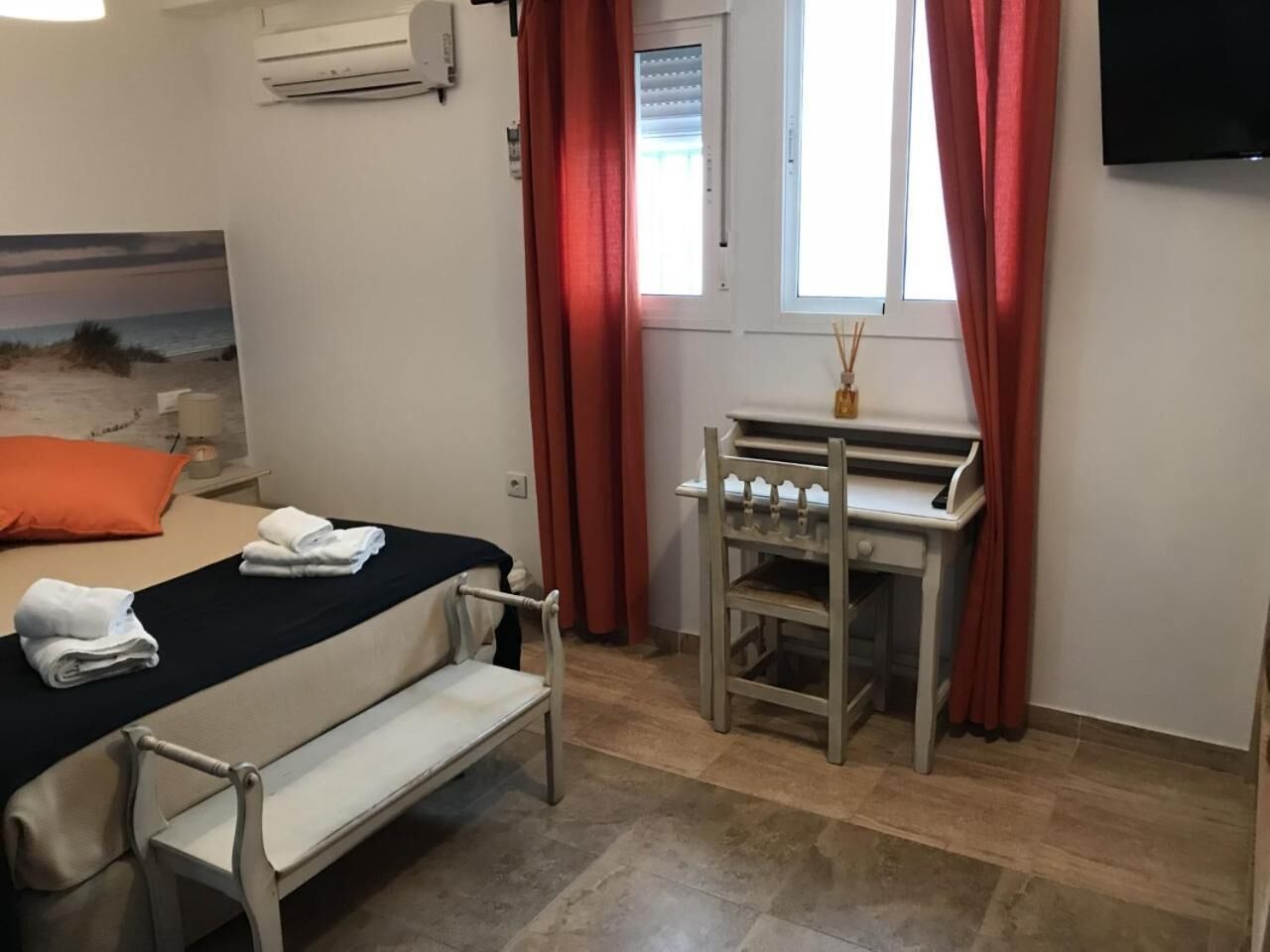 Habitación de hotel con cama, una banca, una mesita con una vela y una ventana con cortinas rojas.