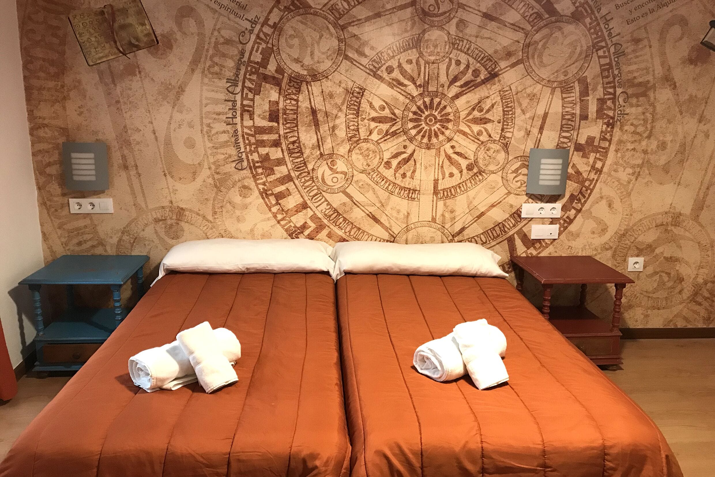 Un lit double avec une literie orange, deux oreillers et deux serviettes enroulées posées sur le lit. Il y a deux tables de chevet, une bleue et une marron, chacune avec une lampe. Le mur est orné d’une grande tapisserie aux motifs complexes.
