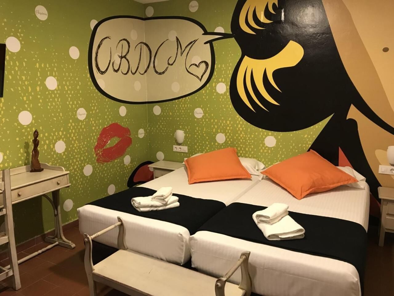 Quarto de hotel com cama, duas almofadas, mesa de cabeceira, escrivaninha e uma cadeira.