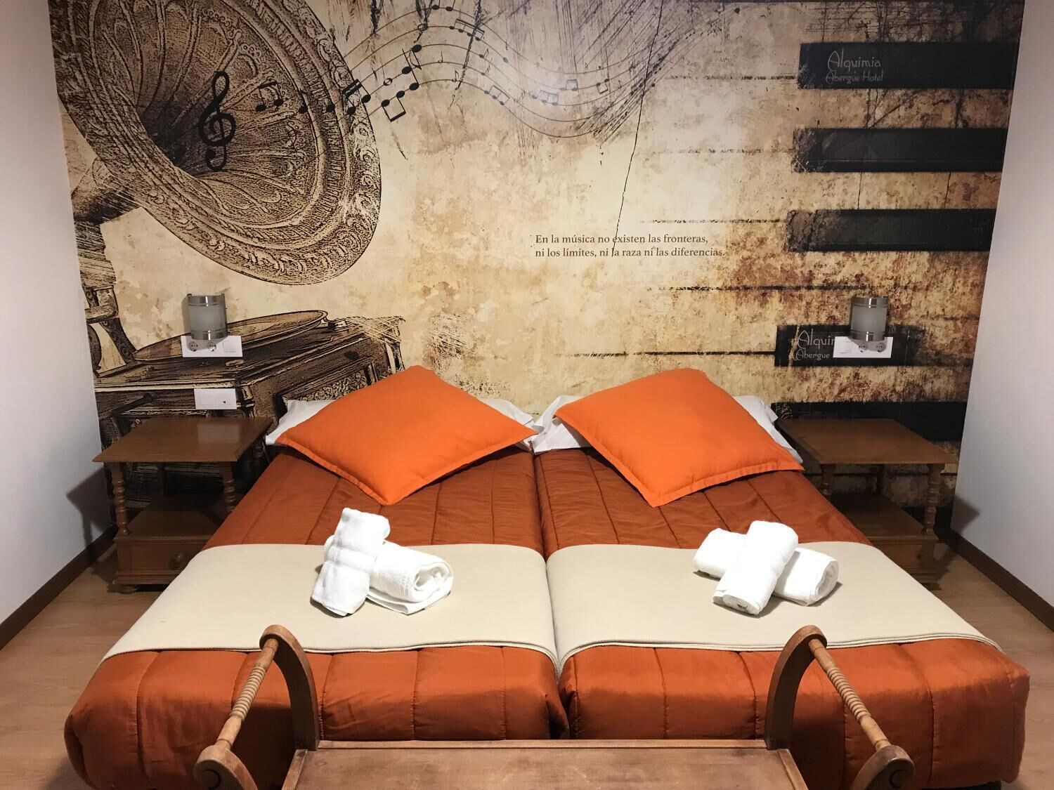 A double room with two beds, each with an orange headboard and pillows, a wooden headboard, and a wall mural with a musical theme.