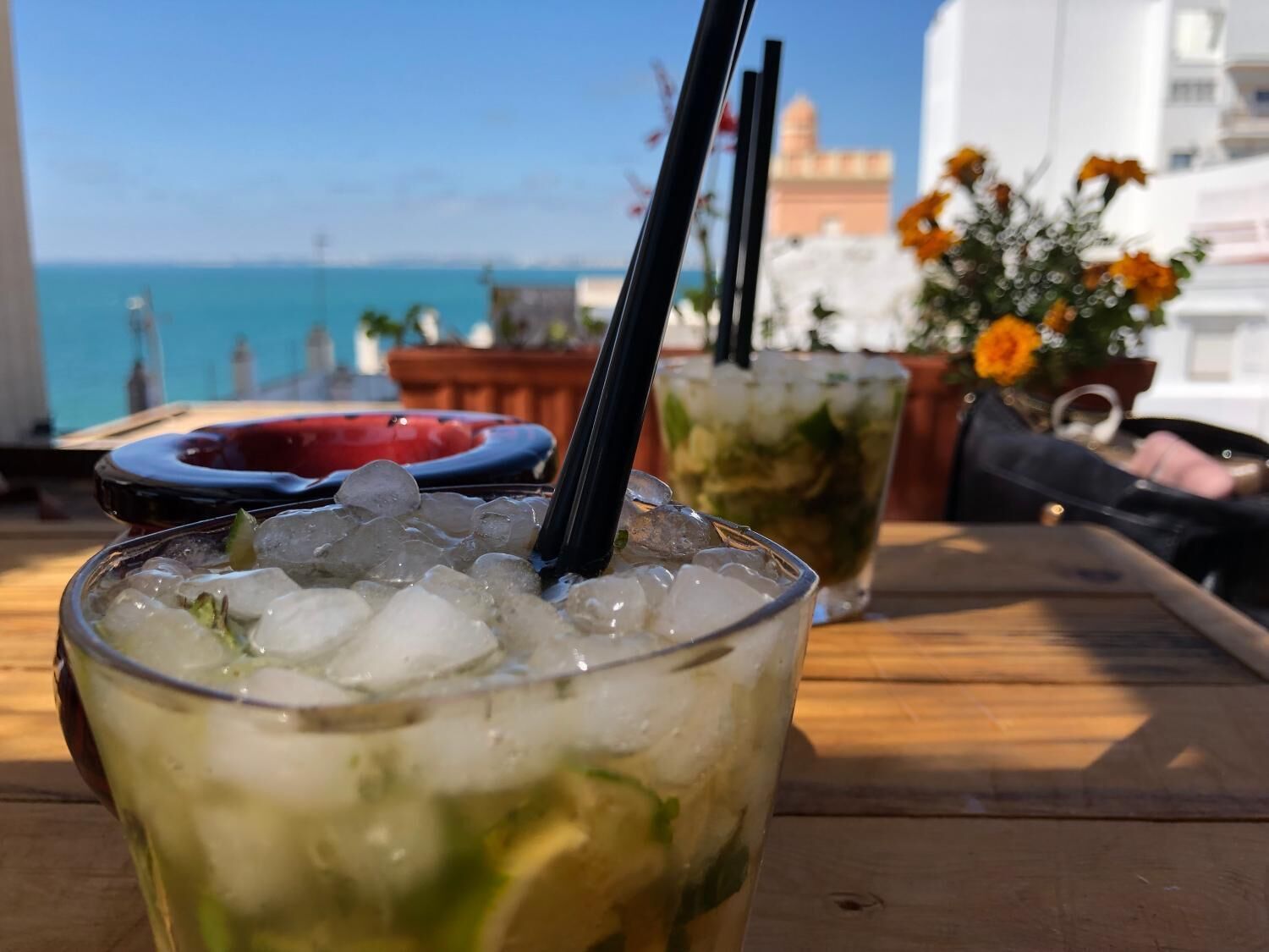 A glass with ice and a straw, a second glass with a drink, and a view of the sea and buildings in the background.