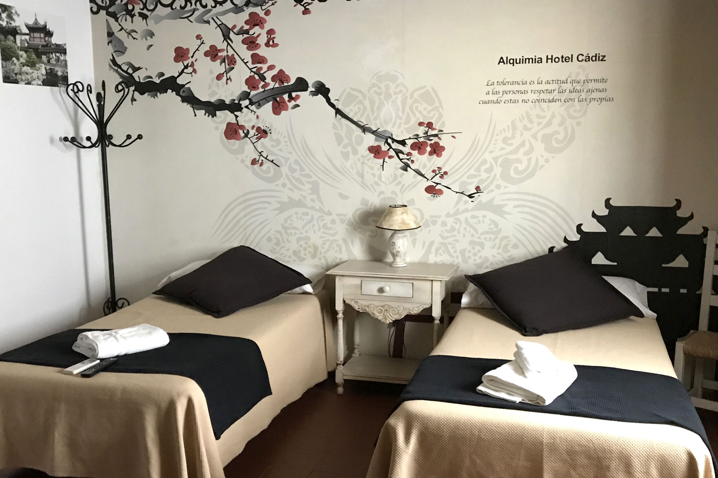 A hotel room with two beds, each with a black and beige bedspread, a small bedside table with a lamp, and a decorative wall mural.