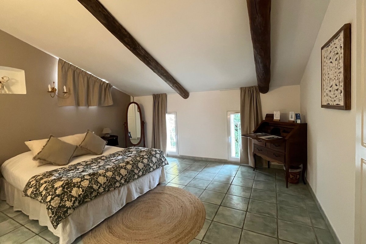 Een slaapkamer met een bed, een bureau en een stoel. Er is een raam met gordijnen, een spiegel en een kunstwerk aan de muur.