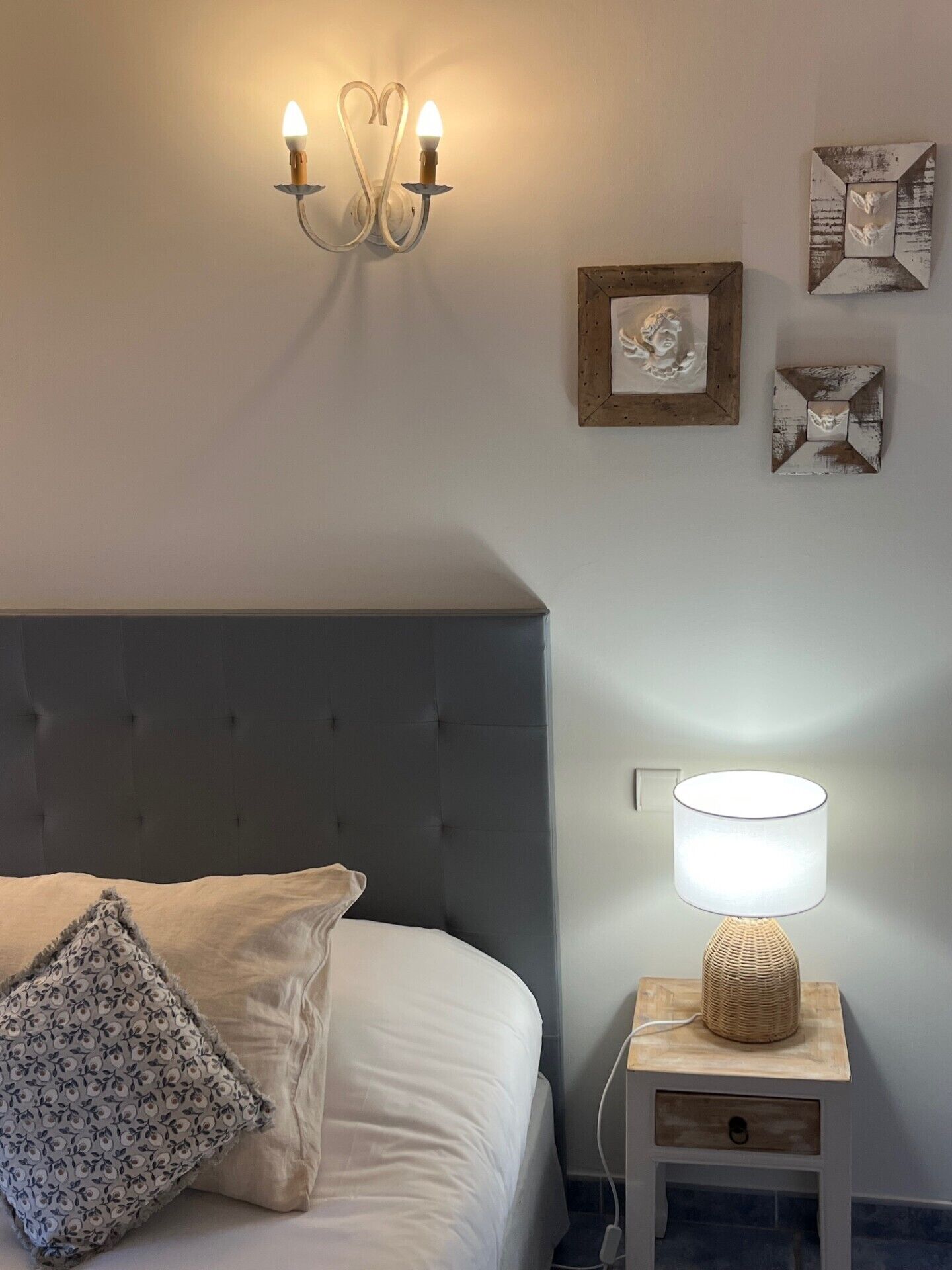 Un lit bien fait, avec une tête de lit grise, une lampe de chevet et une petite table de chevet également dotée d’une lampe. Des appliques murales et des tableaux encadrés ornent le mur.