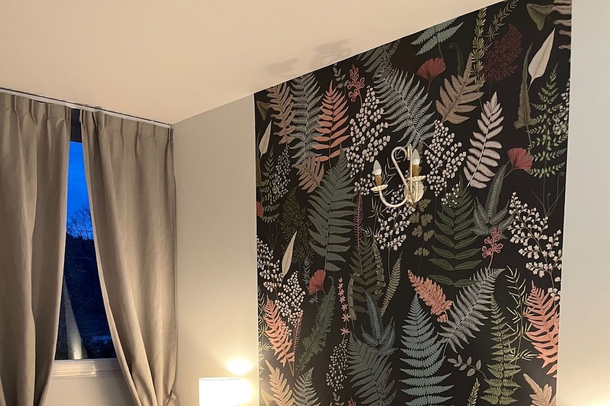 Een slaapkamer met een bed, gordijnen, een nachtkastje, een lamp en een wand met botanisch behang.
