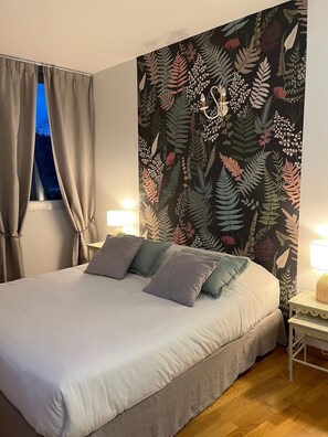 Un dormitorio con cama, cortinas, una mesita de noche, una lámpara y un papel tapiz con motivos botánicos.