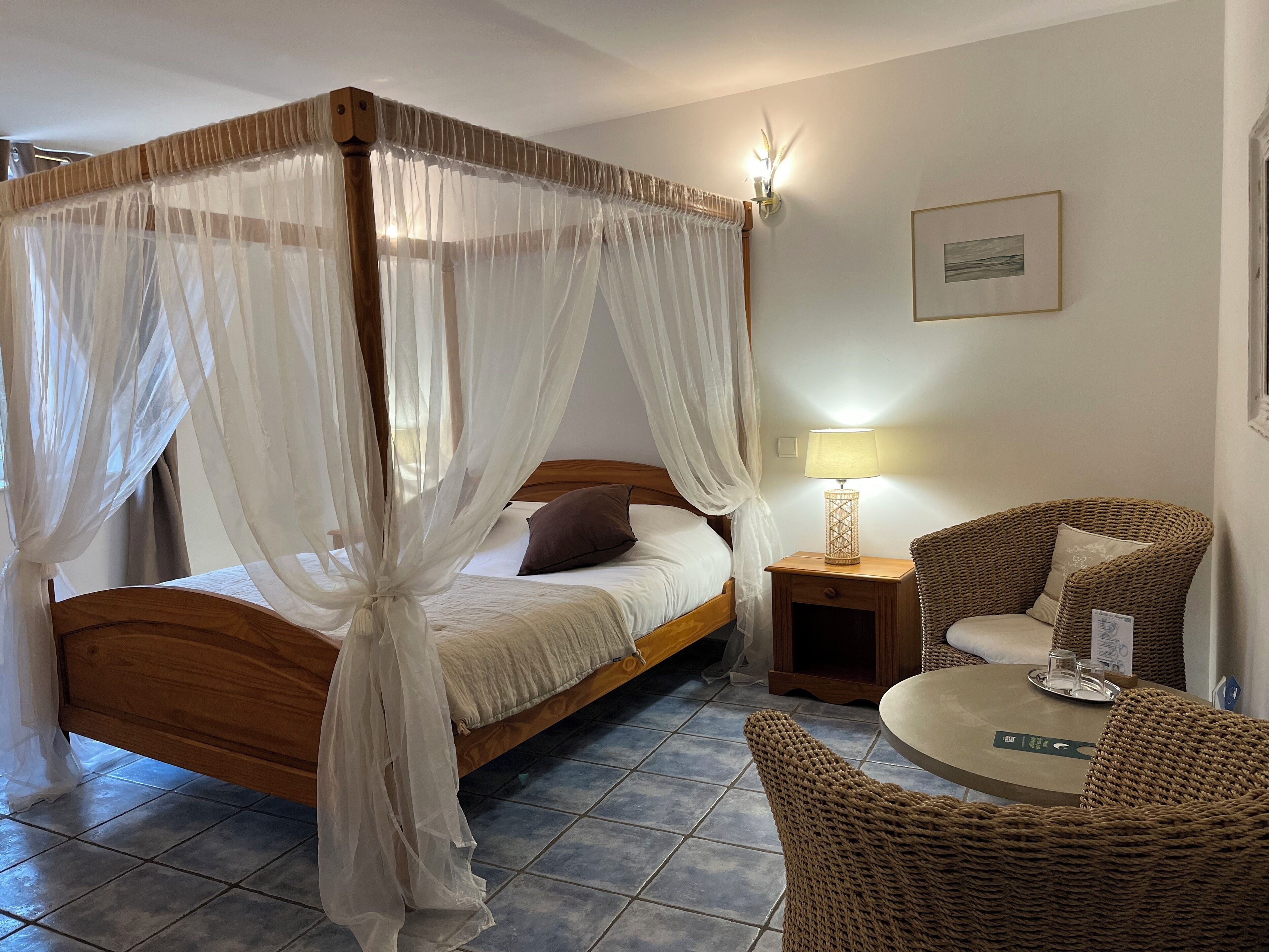 A bedroom with a four-poster bed, a bedside table with a lamp, a wicker chair, and a small table with a tray.