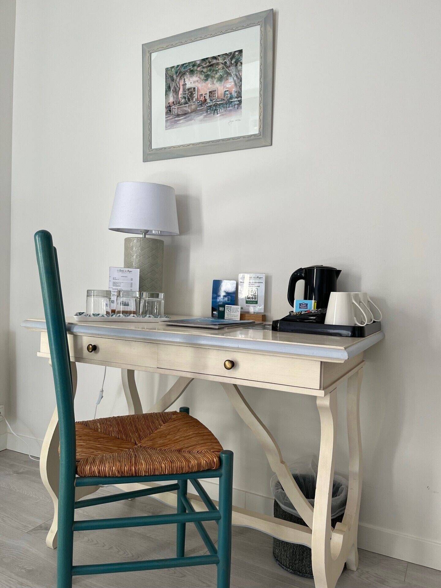A small, neatly arranged workspace with a desk, chair, lamp, coffee maker, and a framed picture on the wall.