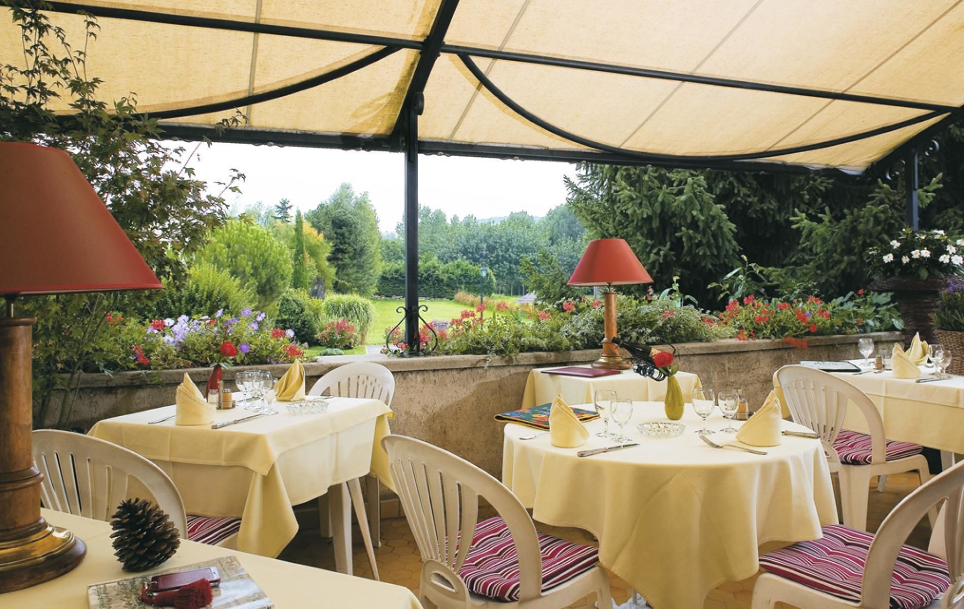 An outdoor dining area with tables set for two, covered by a canopy, and surrounded by lush greenery and flowers.