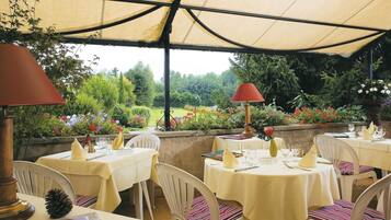 An outdoor dining area with tables set for two, covered by a canopy, and surrounded by lush greenery and flowers.