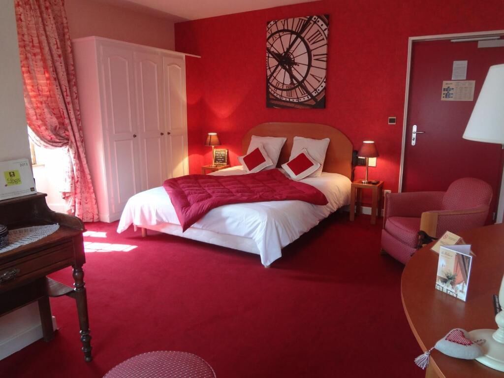 A red-themed hotel room with a bed, a desk, a chair, and a clock on the wall.