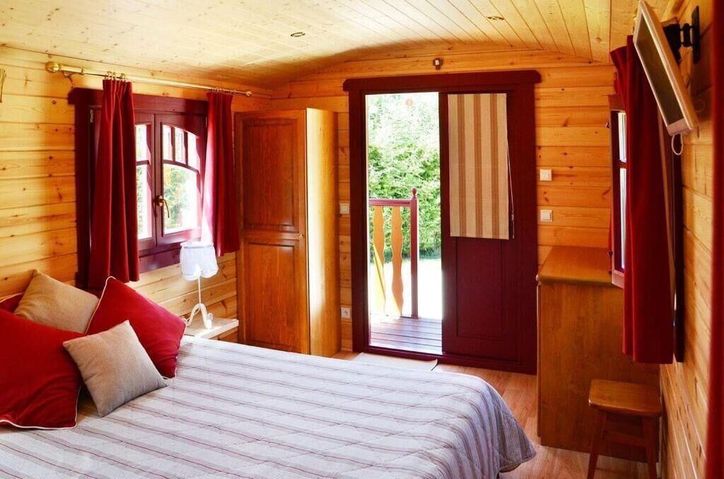 Un dormitori de cabana de fusta amb un llit, cortines vermelles, una taula petita i un televisor fixat a la paret.
