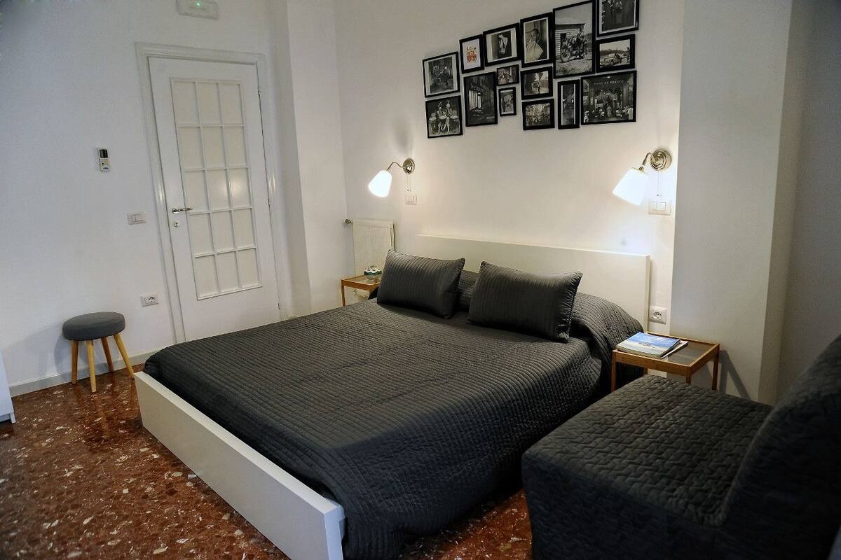 A bedroom with a bed, two bedside tables, a chair, a door, and a wall with framed pictures.