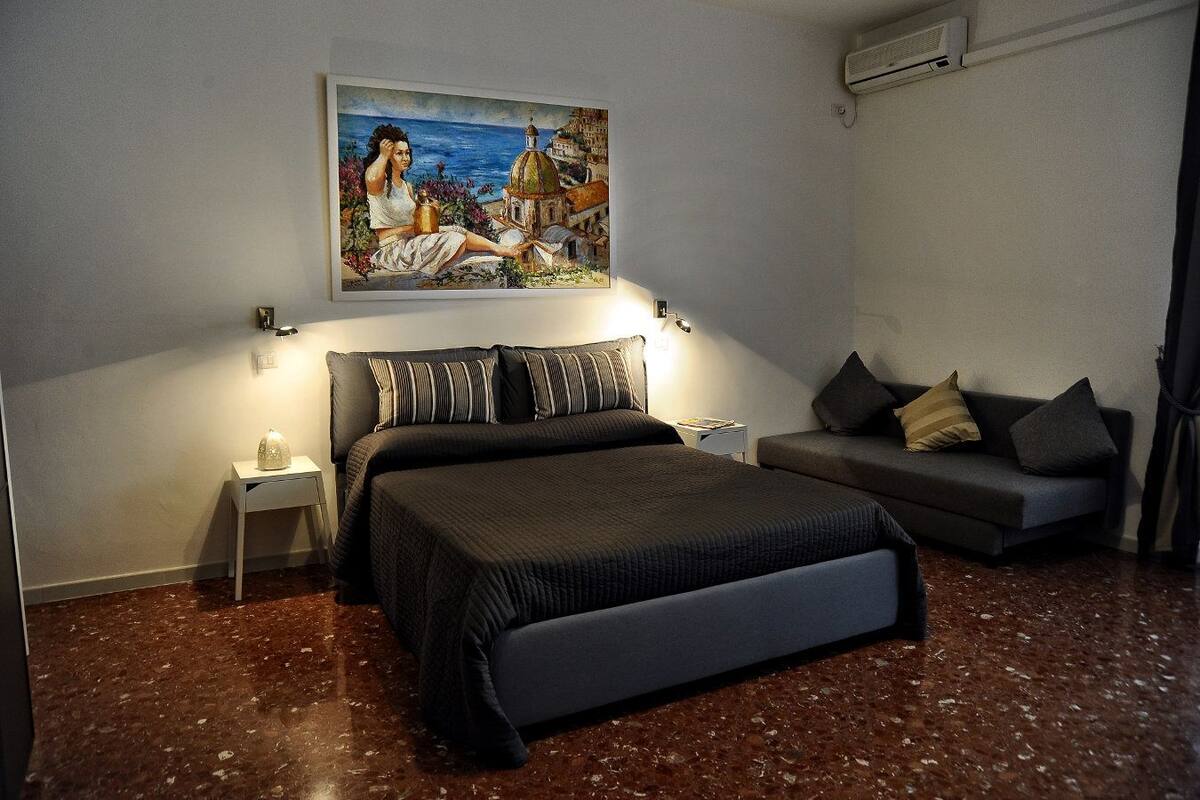 A bedroom with a bed, a bedside table, a nightstand, a sofa, and a painting on the wall.