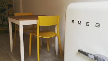 Un comedor con una mesa de madera y una silla amarilla, un refrigerador blanco con la marca 'SMEG' y un cuadro en la pared.
