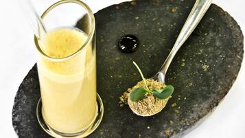 A glass of yellow liquid on a black plate with a spoonful of green leaves and a drop of black liquid.