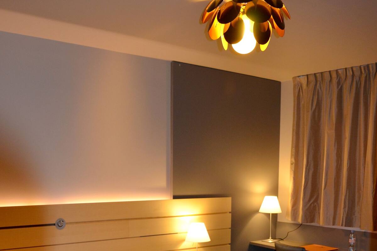A hotel room with a large bed, two bedside lamps, a desk with a chair, a ceiling light fixture, and a window with curtains.