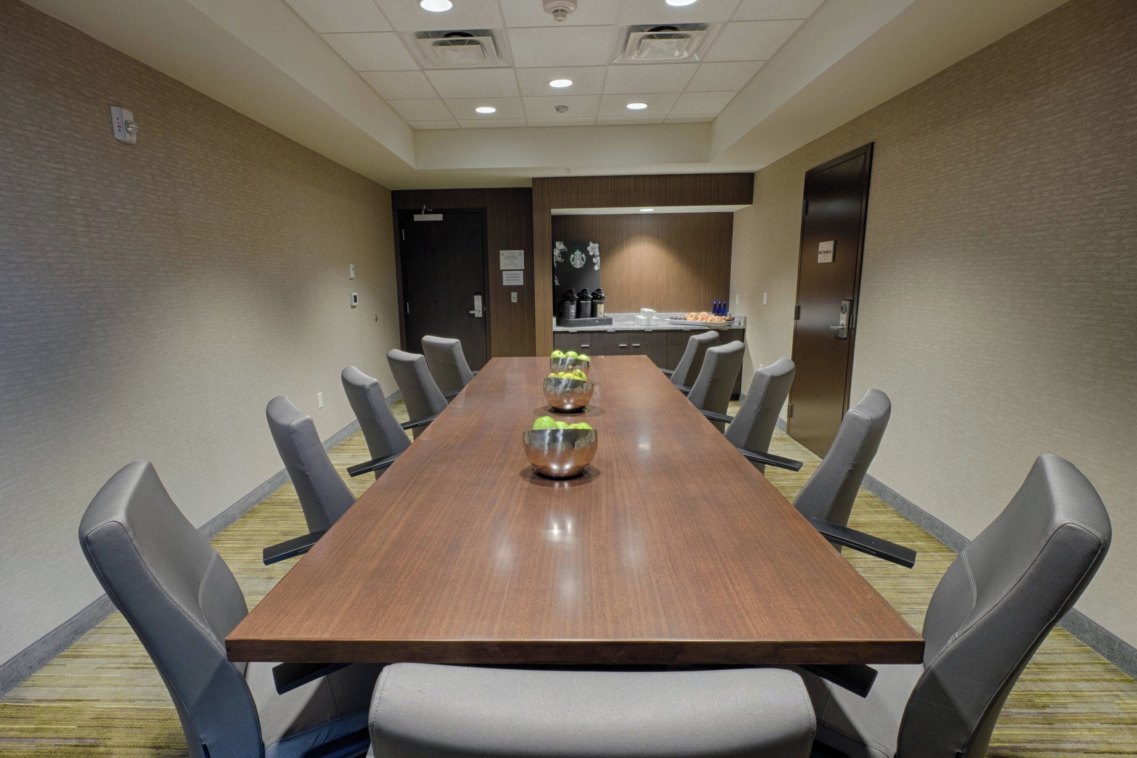 A conference room with a long wooden table, grey upholstered chairs, and a kitchen area in the background.