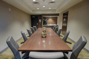 A conference room with a long wooden table, grey upholstered chairs, and a kitchen area in the background.