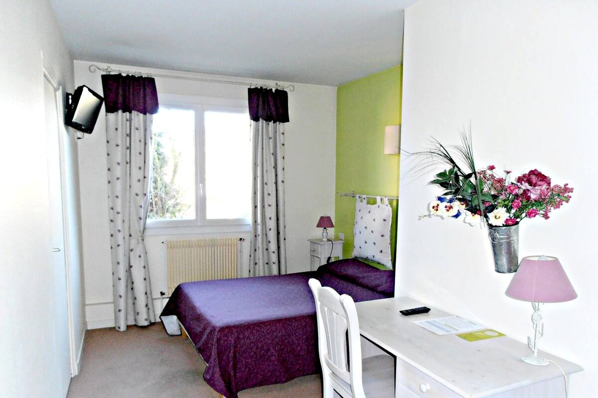 A bedroom with a bed, a desk, a chair, a lamp, a nightstand, and a window with curtains.